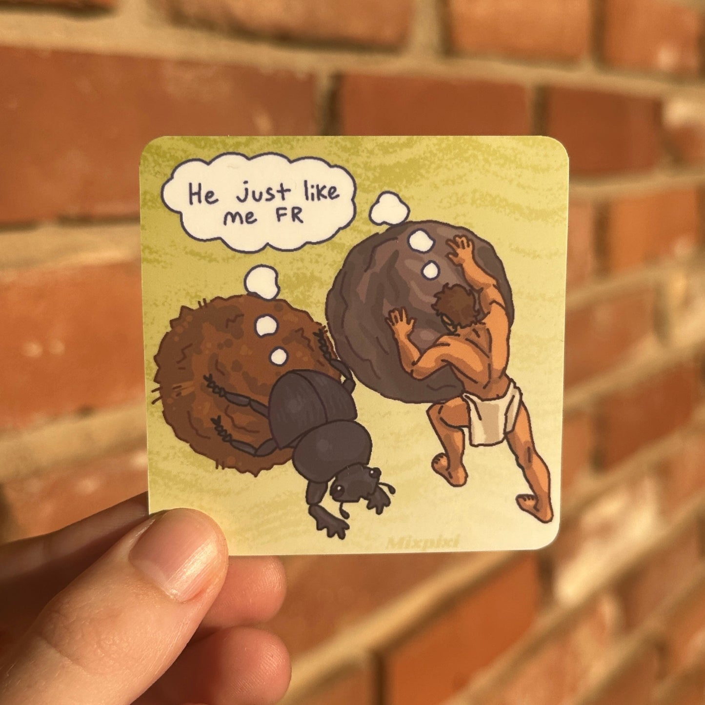 A hand holds a square illustrated sticker in front of a blurred brick wall. The artwork shows a cartoon figure pushing a large dark boulder, with a speech bubble reading 'He just like me FR'.