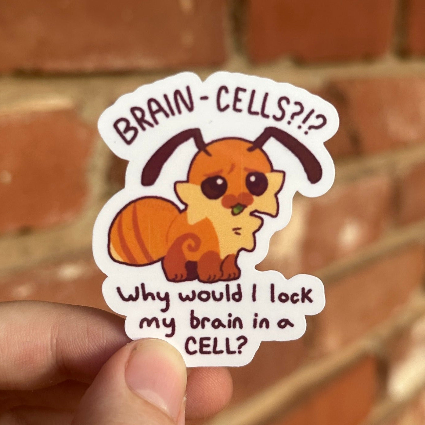 A hand holds a die-cut sticker featuring a cartoon leaf cutter ant with the text 'BRAIN-CELLS??' and 'Why would I lock my brain in a CELL?' against a blurred red brick wall background.
