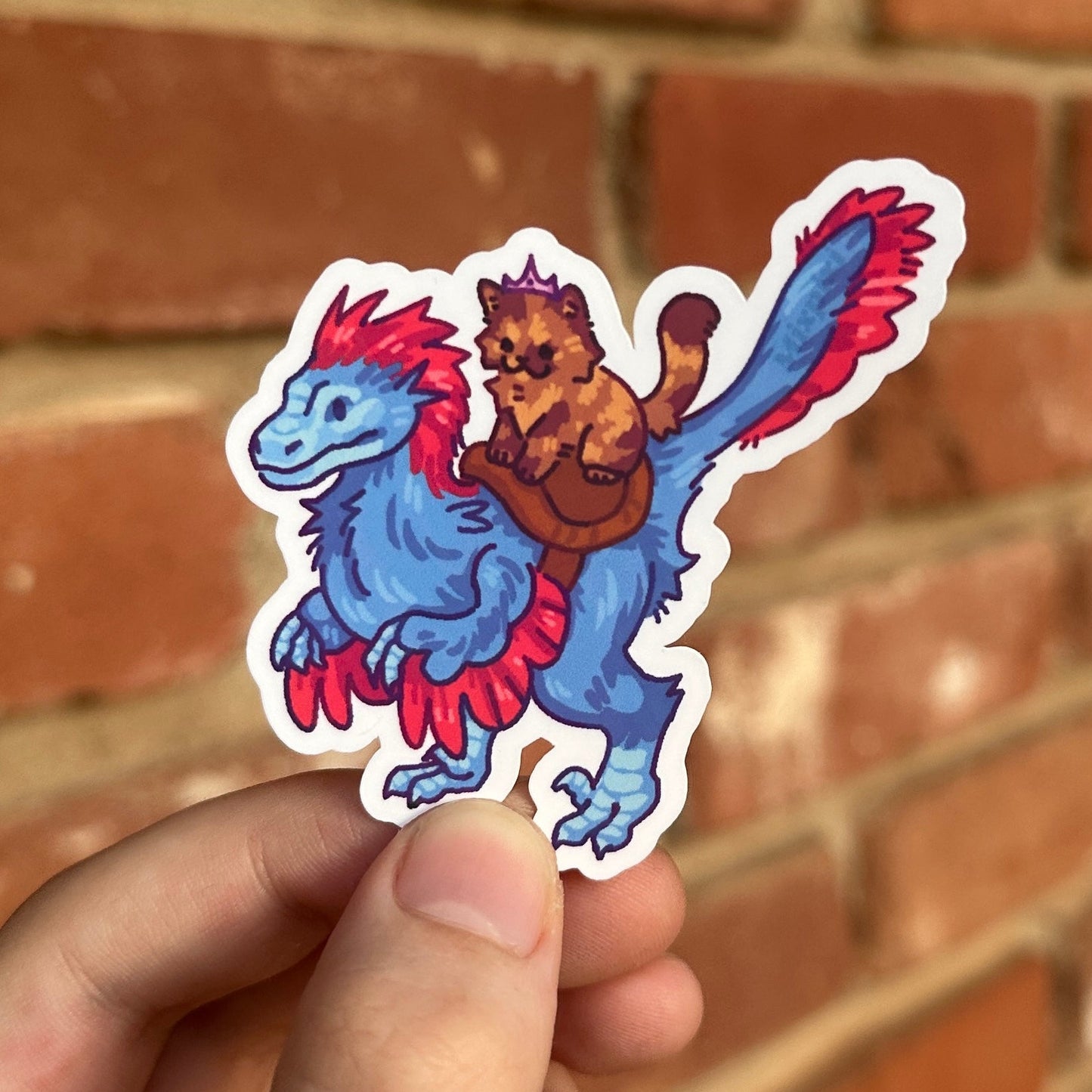 A hand holds a die-cut sticker featuring a blue dinosaur-like creature with red accents and a small brown cat riding on its back, set against a blurred red brick wall background.
