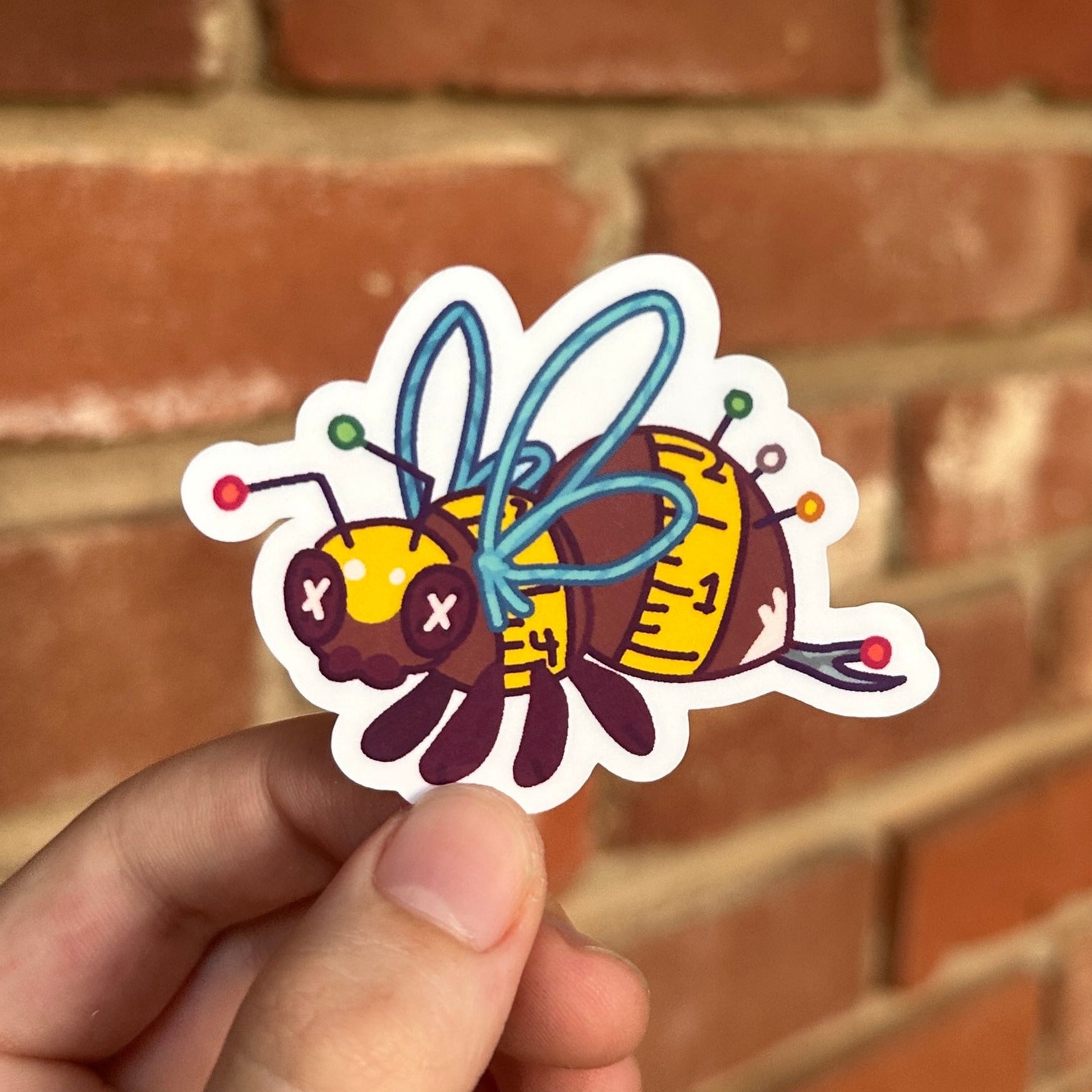 A hand holds a die-cut sticker of a cartoon wasp with blue wings and a yellow-and-black striped body, set against a blurred red brick wall background.