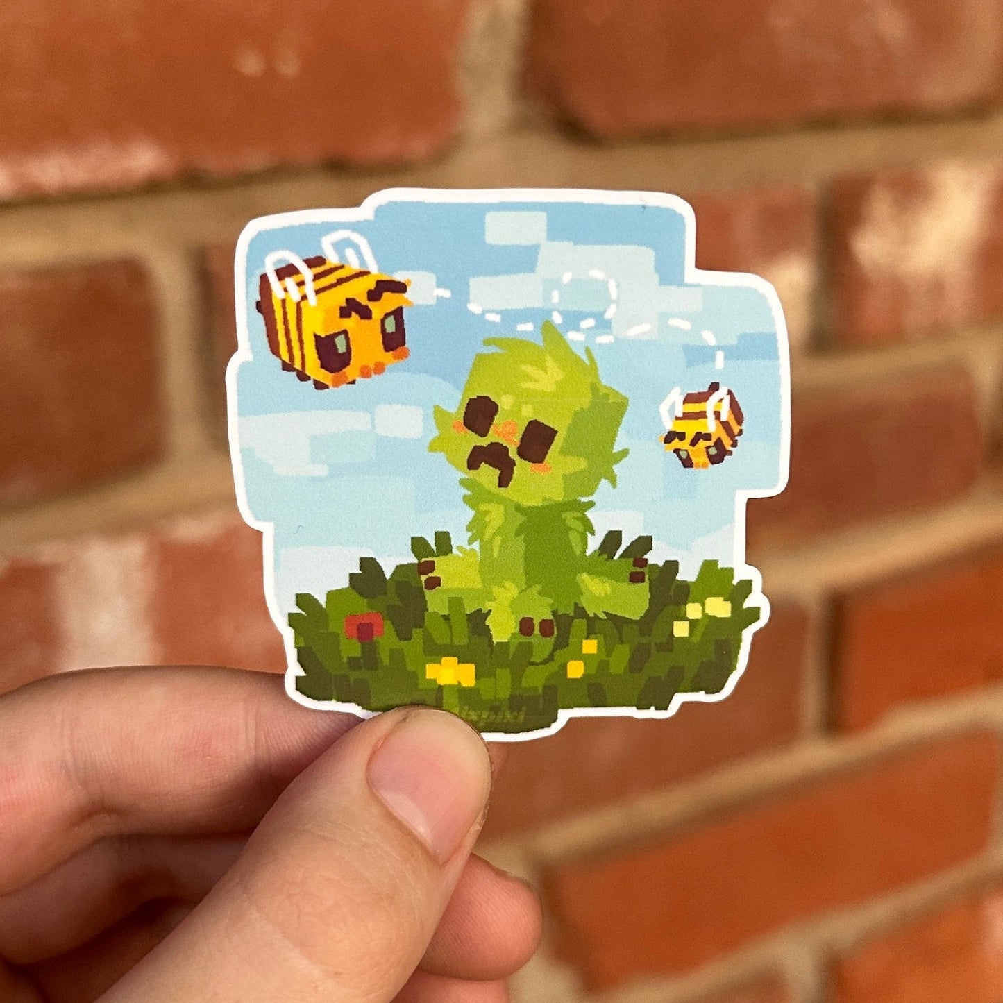 A hand holding a die-cut sticker featuring a pixel-art green guy with bees against a blue sky, set against a blurred red brick wall background.