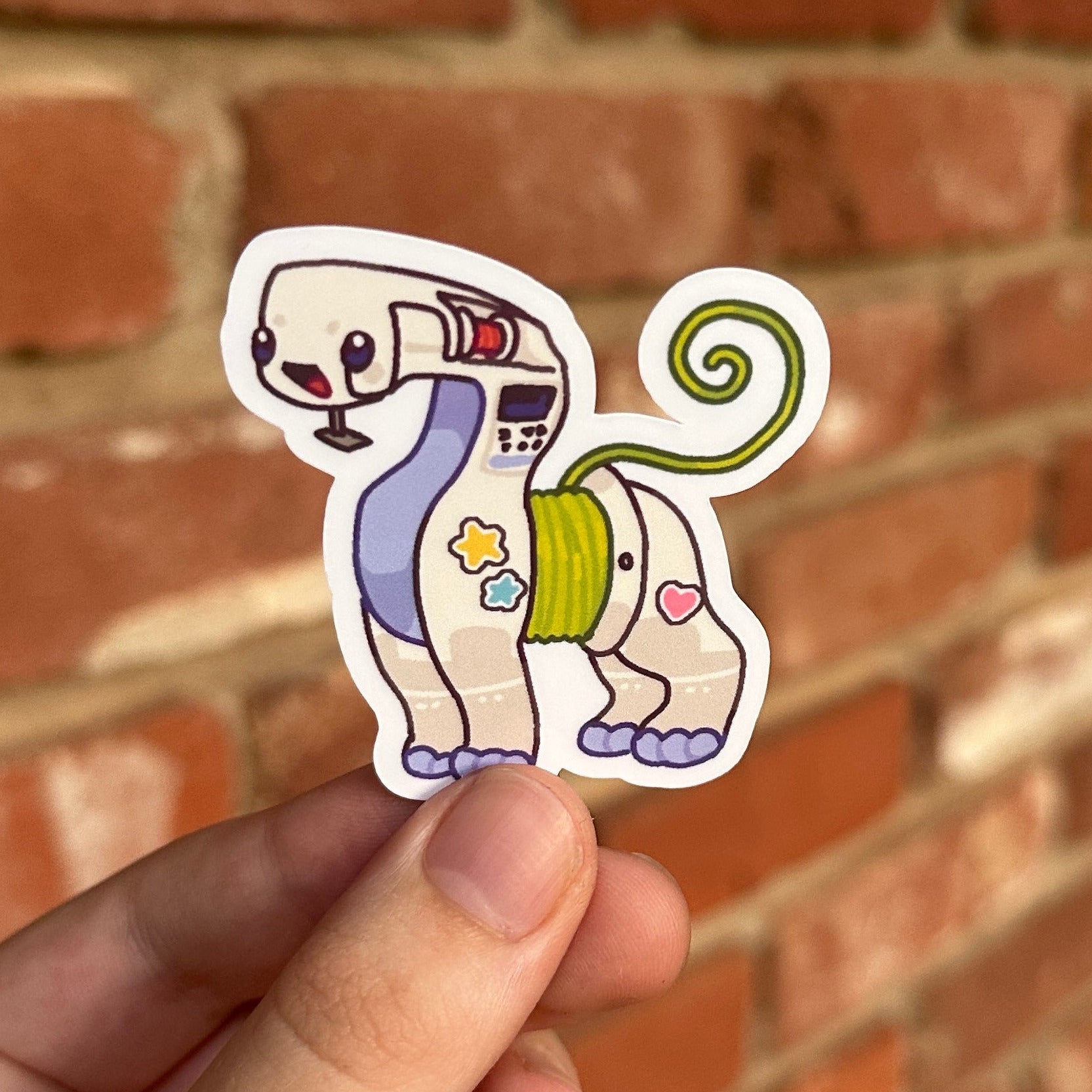 A hand holding a die-cut sticker of a personified sewing machine, set against a blurred red brick wall background.