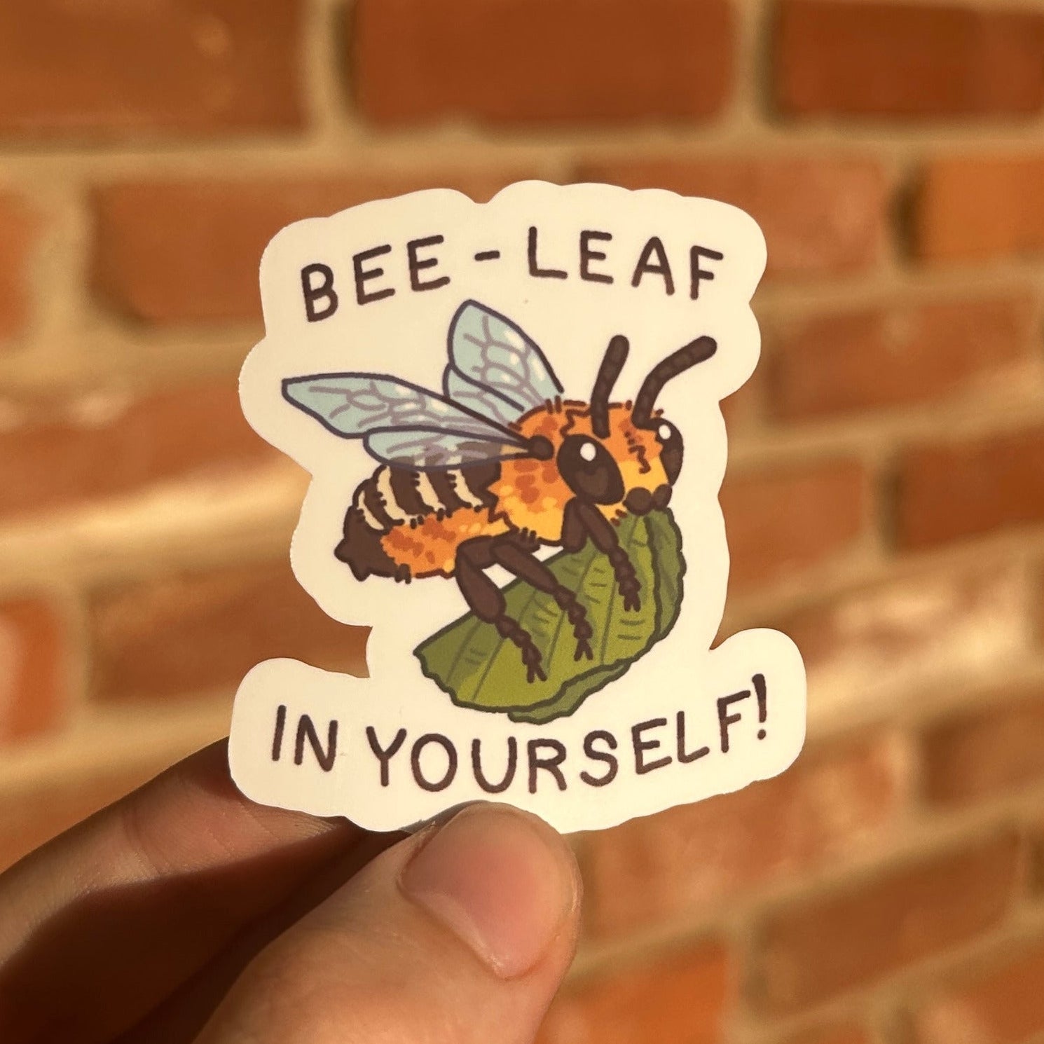 A hand holds a die-cut sticker featuring an illustrated bee on a leaf with the text 'BEE-LEAF IN YOURSELF!' against a blurred red brick wall background.
